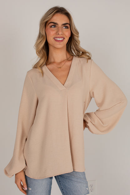 A woman wearing a light beige long-sleeve blouse with a V-neckline and balloon sleeves, paired with blue jeans, standing against a plain light gray background.