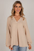 A woman wearing a beige long-sleeve V-neck blouse with a gathered front, paired with light blue jeans, against a solid light gray studio background.
