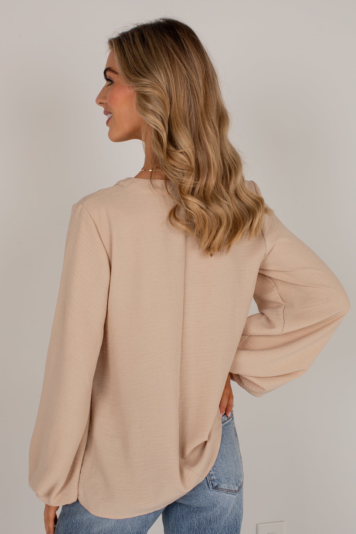 A woman shown from the back wearing a light beige long-sleeve blouse with balloon sleeves and blue jeans, standing against a plain light gray wall.