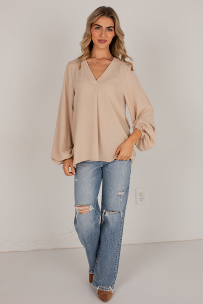 A woman modeling a beige long-sleeve blouse with a V-neck and balloon sleeves, paired with distressed blue jeans and brown shoes, standing against a plain light gray wall.