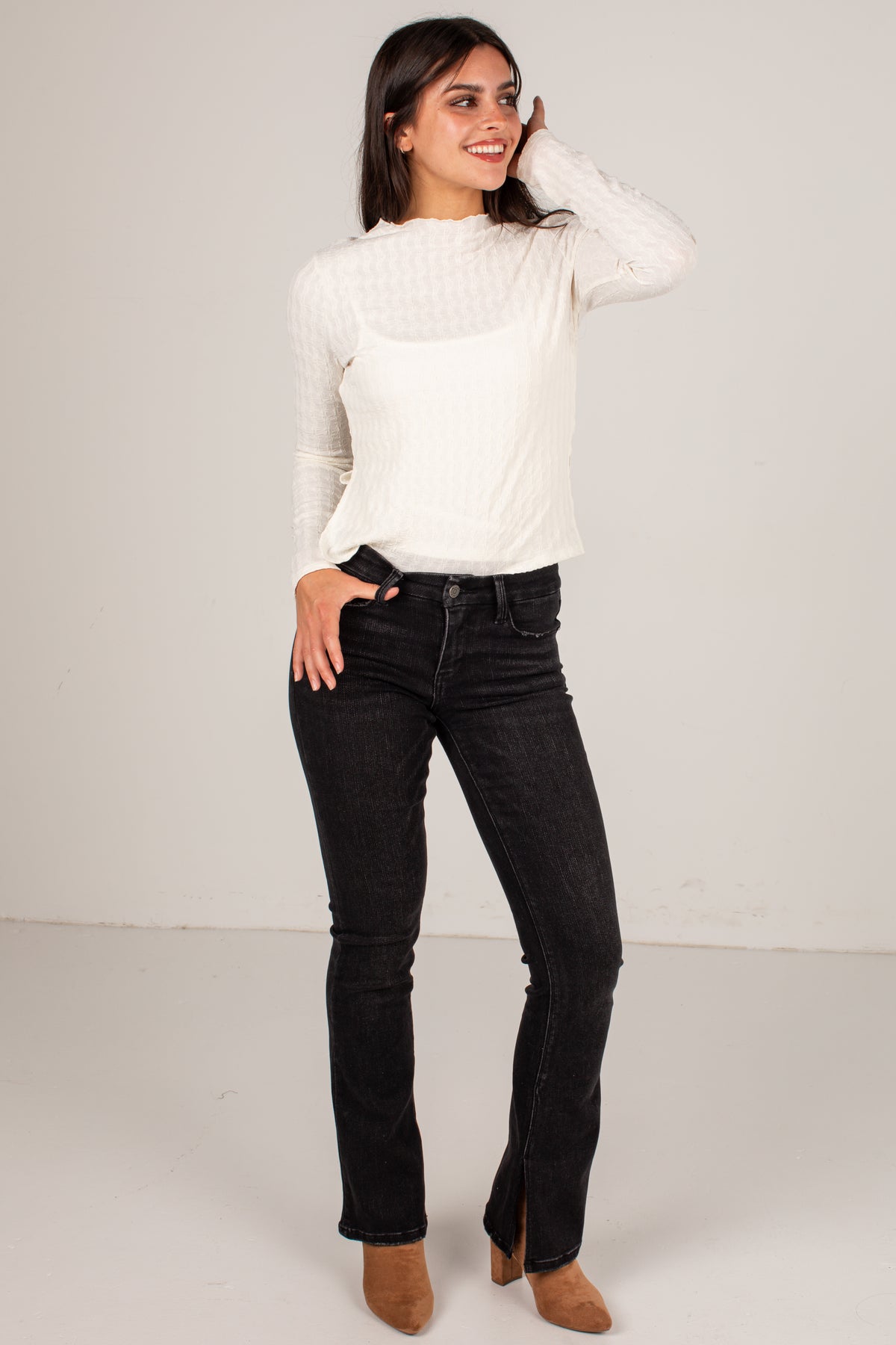 A woman modeling a white long-sleeve knit top and black flared jeans with brown ankle boots, standing against a plain light gray studio background.