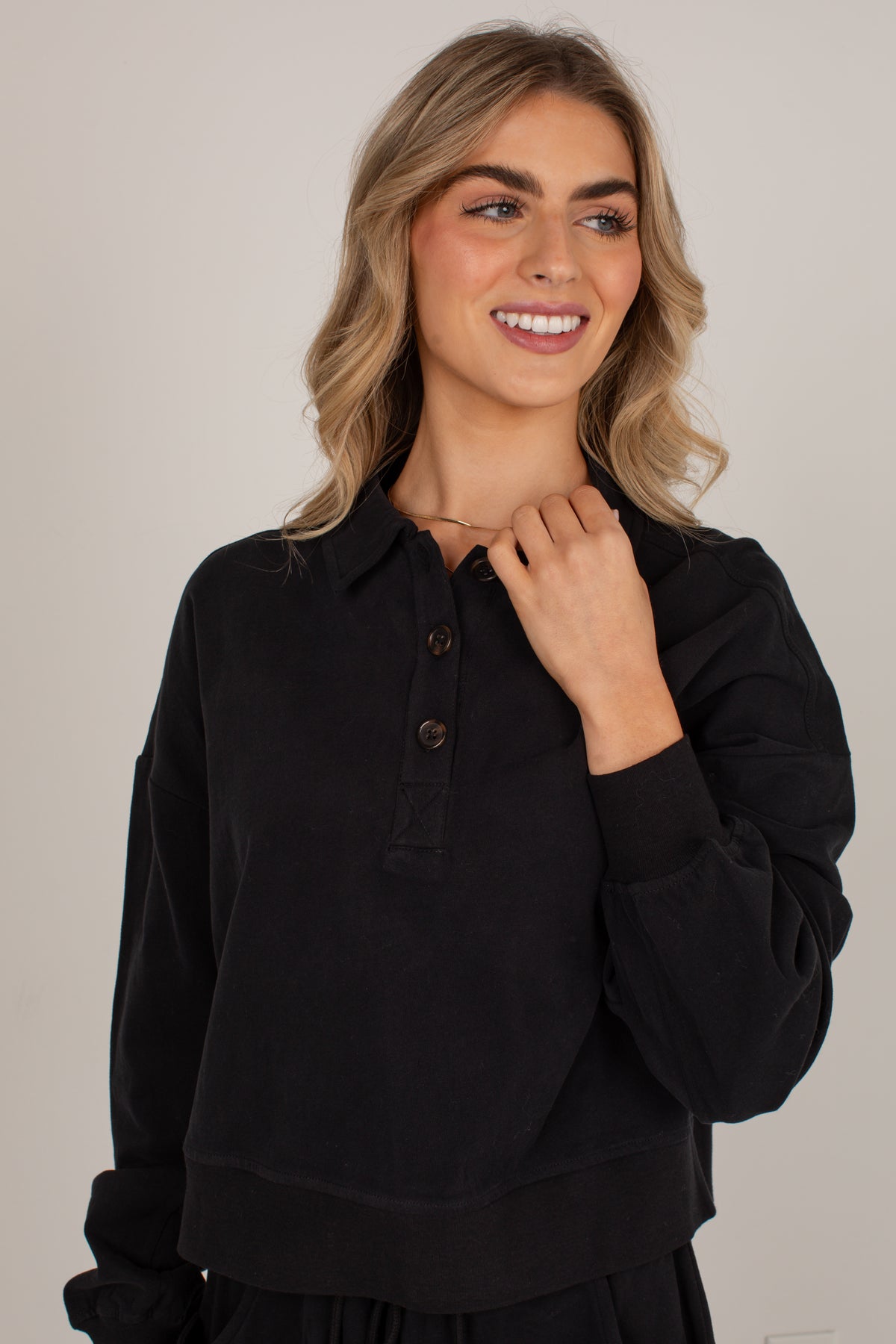 A woman with blonde hair wearing a black long-sleeve button-up shirt, posing against a neutral light gray background.