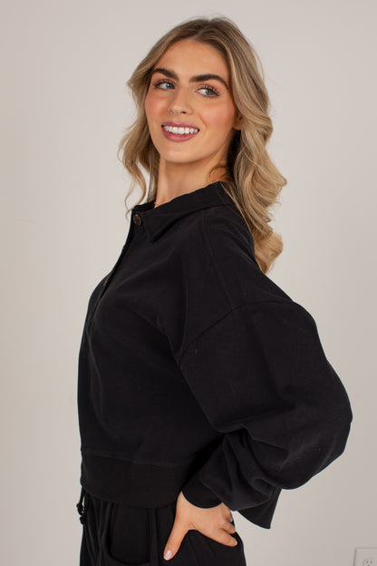 A woman with blonde hair wearing a black cropped hoodie with voluminous balloon sleeves, photographed against a plain light gray studio background.