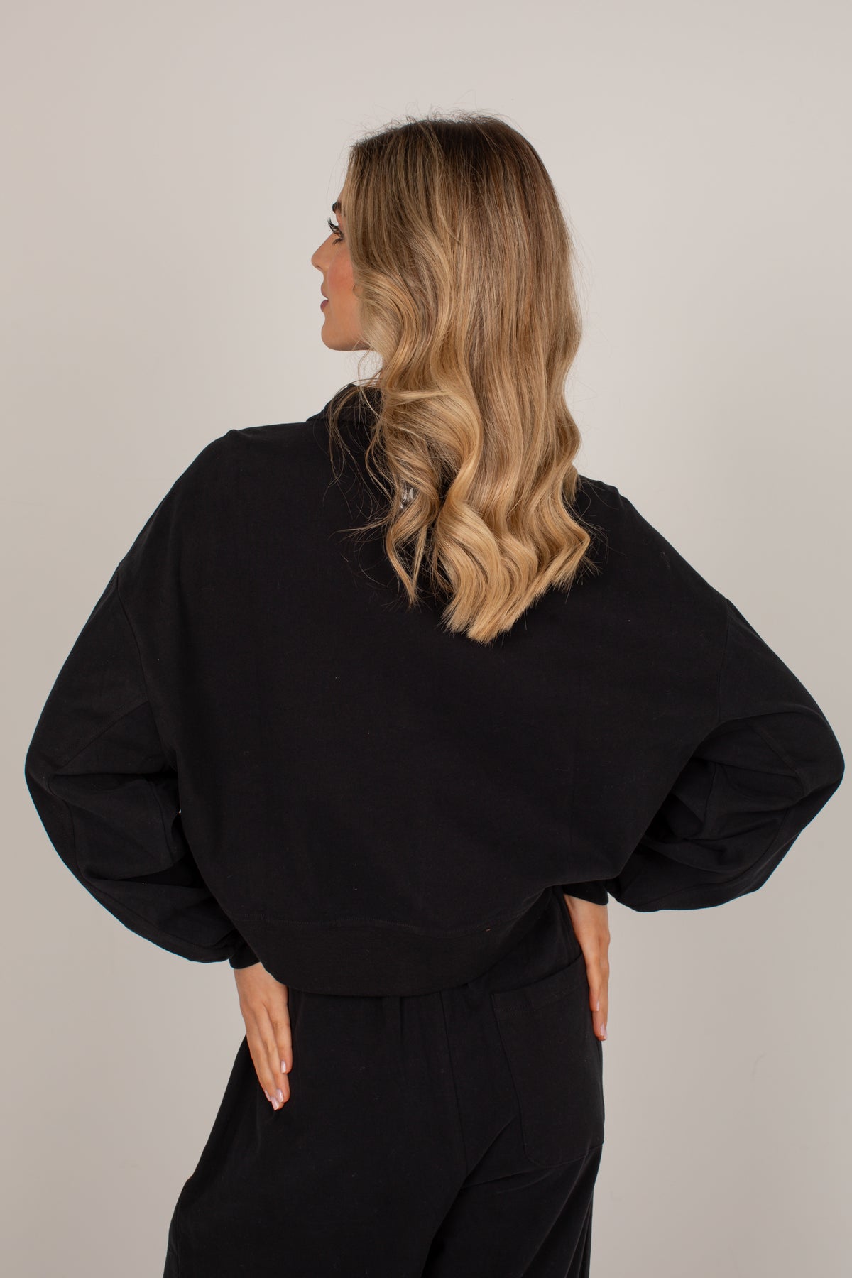 A woman with blonde wavy hair is shown from the back wearing a black oversized long-sleeve top and black pants against a plain light gray studio background.