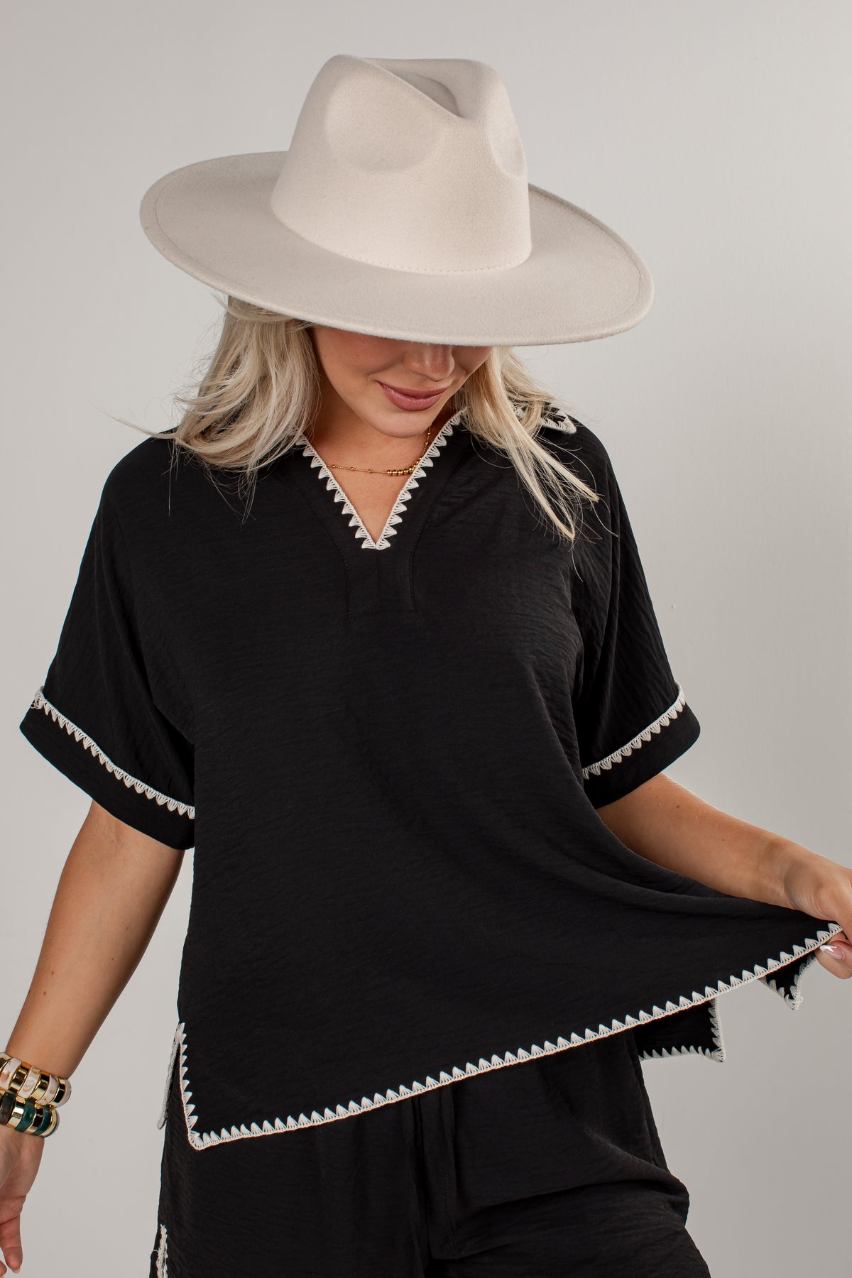 A woman wearing a black short-sleeve top with white decorative trim along the V-neck, sleeve cuffs, and hem, paired with a wide-brimmed cream hat against a light gray studio background.