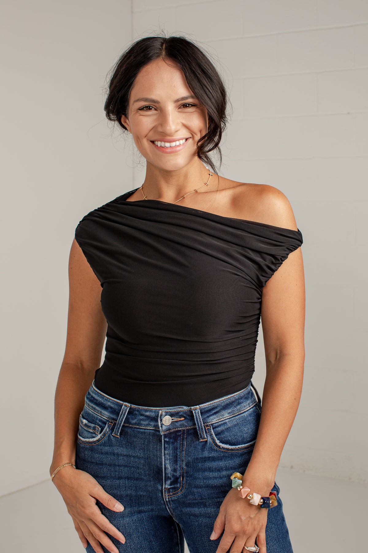 A woman wearing a black off-the-shoulder ruched top and blue jeans, standing against a light gray brick wall.