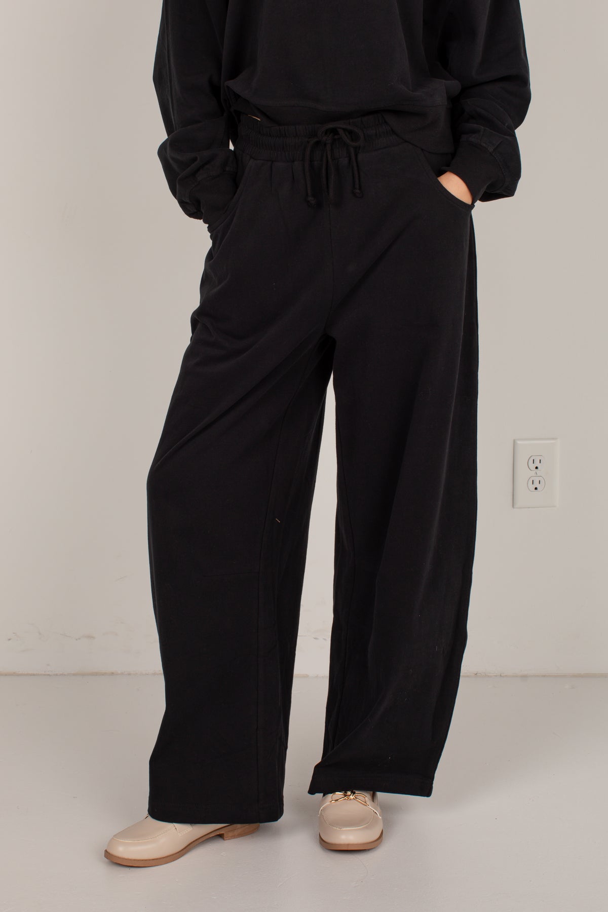 A person wearing loose black wide-leg drawstring pants with a black sweatshirt and beige shoes, standing against a plain light gray wall.