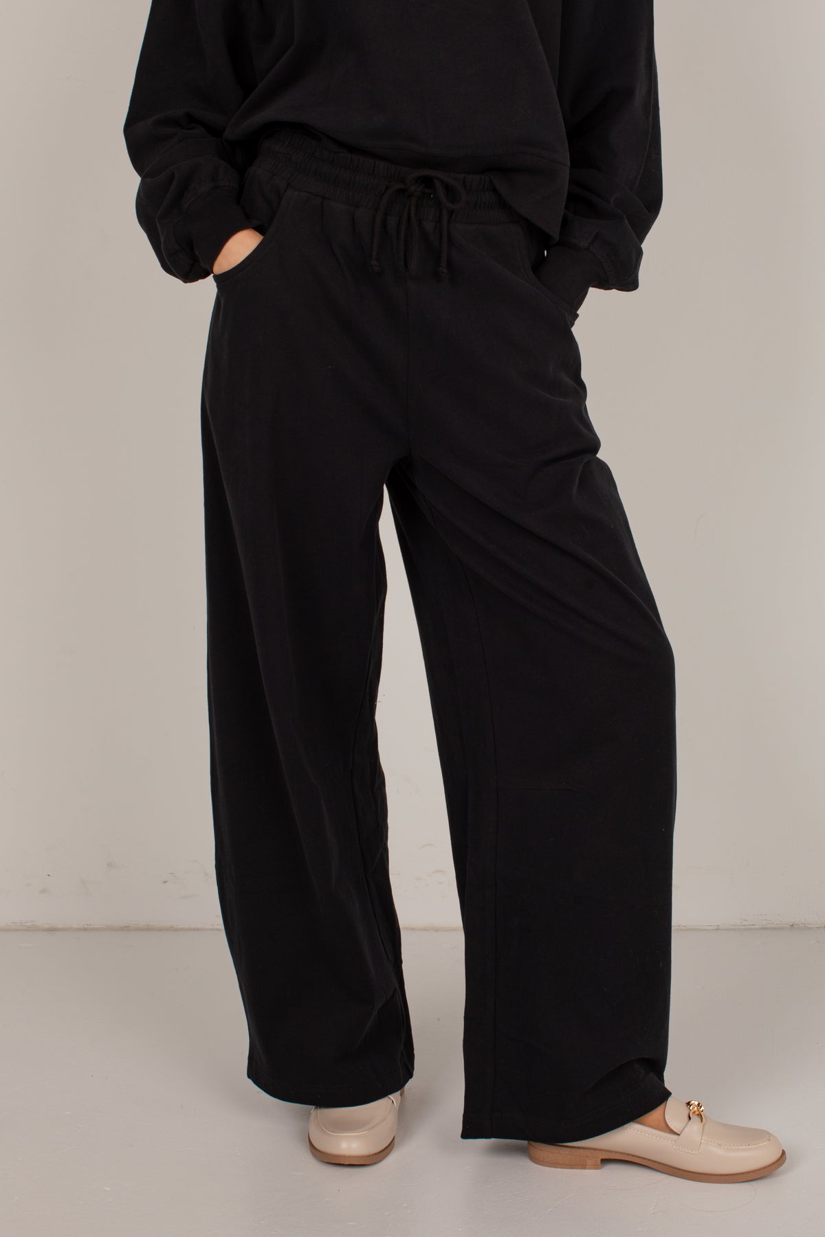 A person wearing loose black wide-leg drawstring pants with a black long-sleeve top and beige loafers, standing against a plain light gray studio background.
