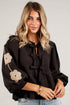 A woman wearing a black long-sleeve blouse with beige floral embroidery on the sleeve and tie-front detailing, paired with light-wash blue jeans, standing against a white paneled wall.