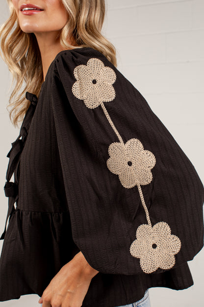 A close-up of a woman wearing a black textured blouse with three large beige crochet flower appliqués down one sleeve, paired with light blue jeans against a white paneled wall.
