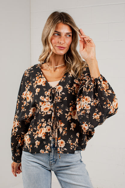 A woman wearing a black long-sleeve blouse with a warm-toned orange and beige floral print, paired with light-wash blue jeans, standing against a white brick wall.