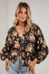 A woman wearing a black long-sleeve blouse with a warm-toned floral print and tie-front detail, paired with light-wash blue jeans, standing against a white paneled wall.