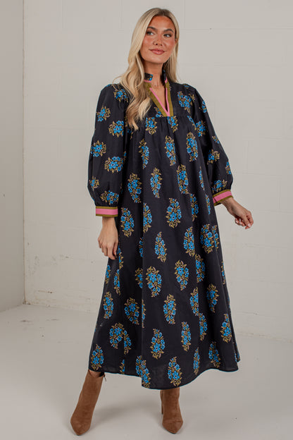 A woman modeling a long-sleeved black maxi dress with a blue and gold floral pattern and pink trim on the cuffs, paired with brown suede boots against a plain white studio background.