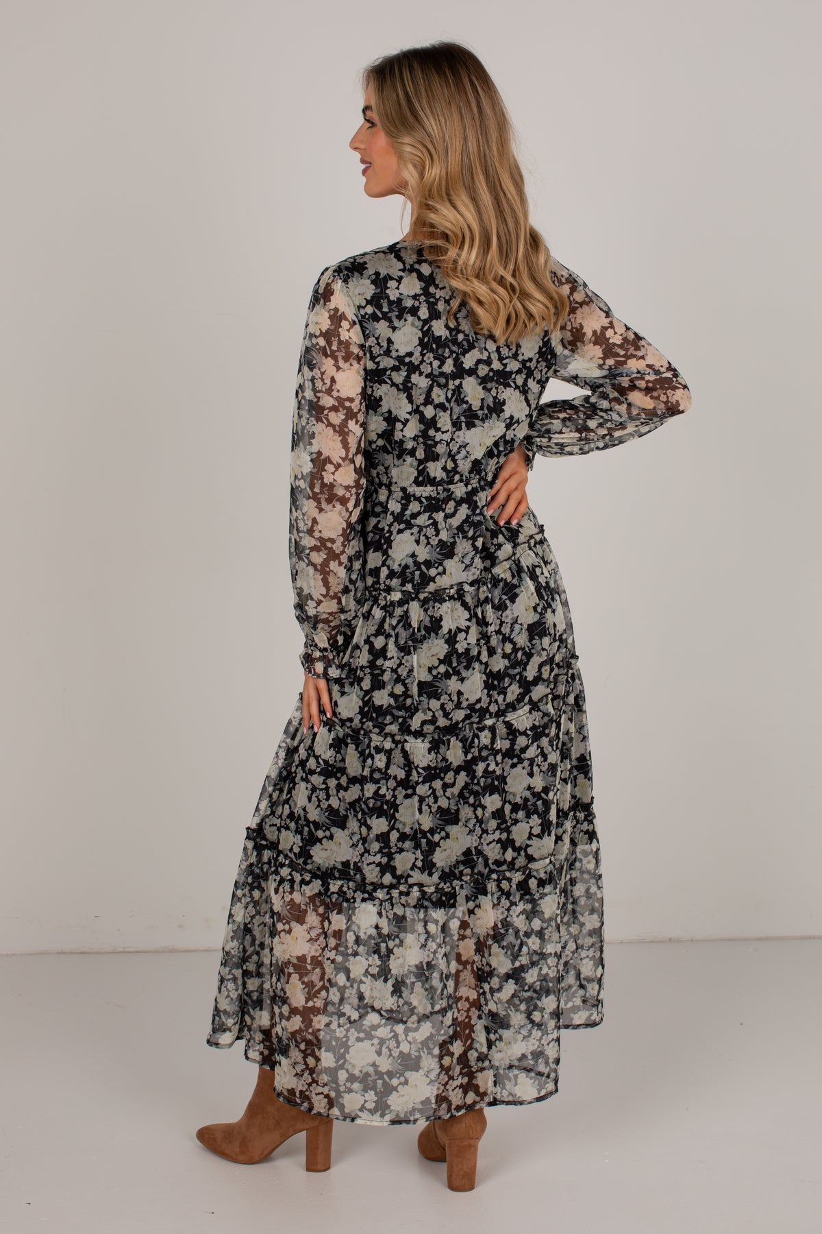 A woman modeling a long-sleeve midi dress with a black, white, and brown floral pattern, shown from a back-three-quarter view against a plain light gray studio background.