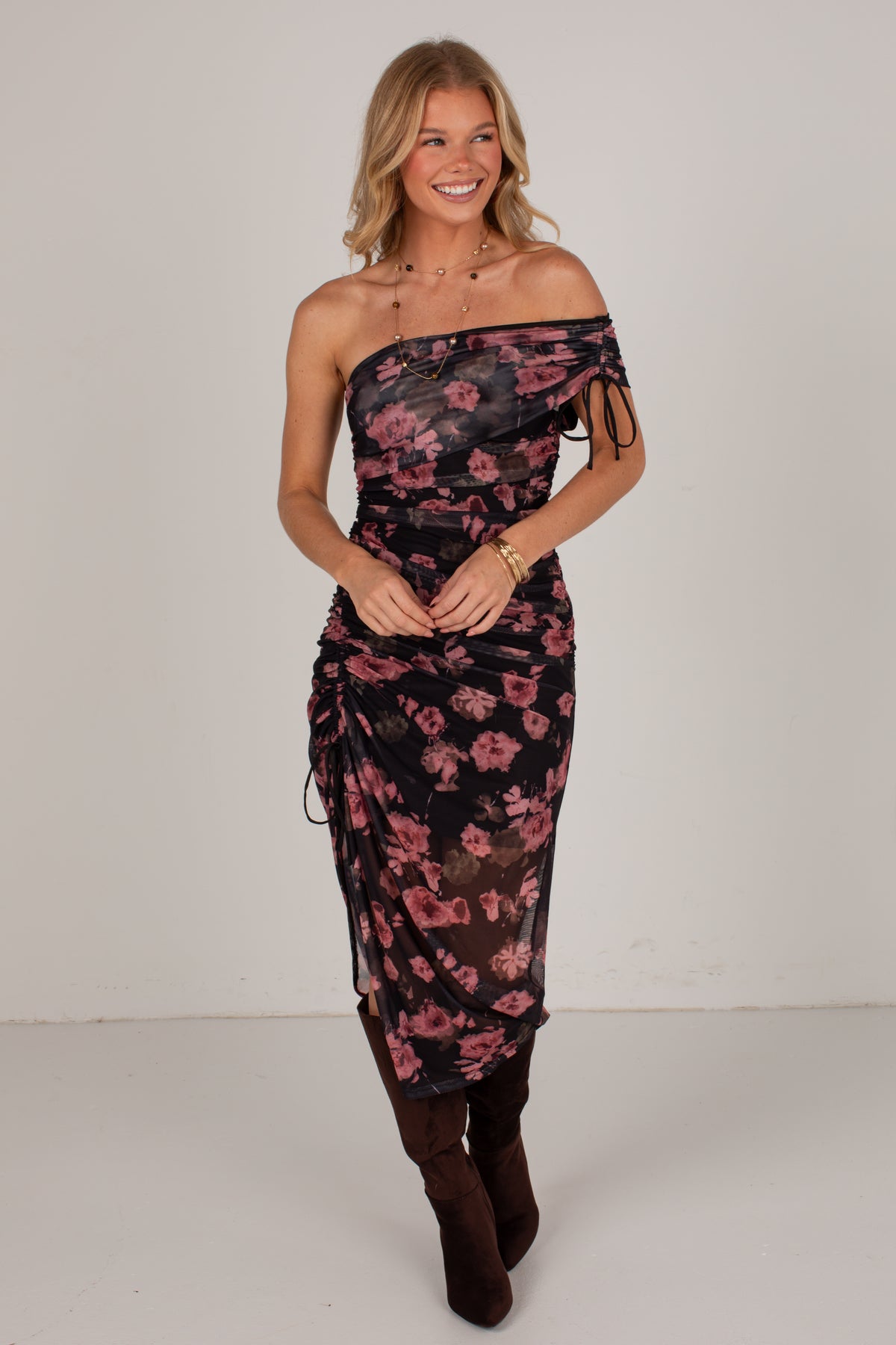 A woman modeling a one-shoulder sheer midi dress with a pink floral print on a black background, paired with dark brown boots, against a plain light gray studio backdrop.
