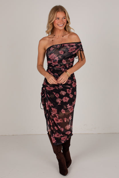A woman modeling a one-shoulder sheer midi dress with a pink floral print on a black background, paired with dark brown boots, against a plain light gray studio backdrop.