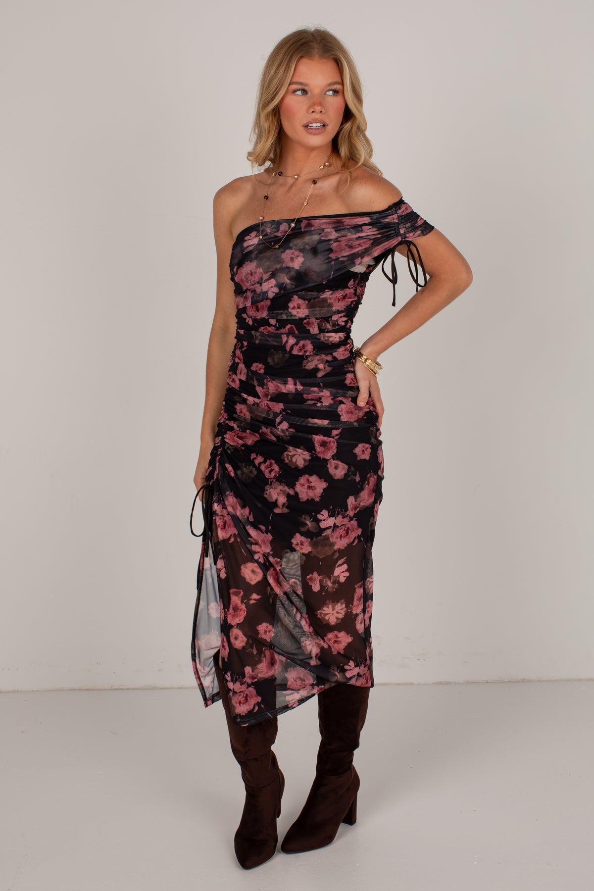 A woman modeling a one-shoulder midi dress with a black base and pink floral print, styled with dark brown knee-high boots against a plain light gray studio background.
