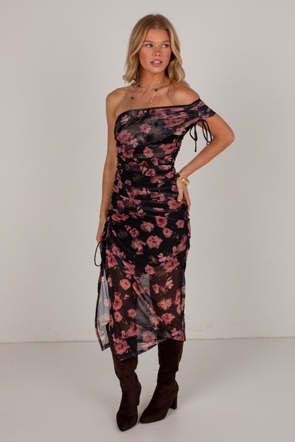 A woman modeling a one-shoulder midi dress with a black base and pink floral print, styled with dark brown knee-high boots against a plain light gray studio background.
