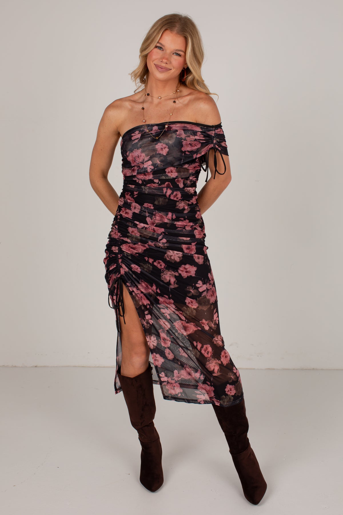 A woman modeling an off-the-shoulder midi dress with a black base and pink floral pattern, paired with dark brown knee-high boots, against a plain light gray studio background.