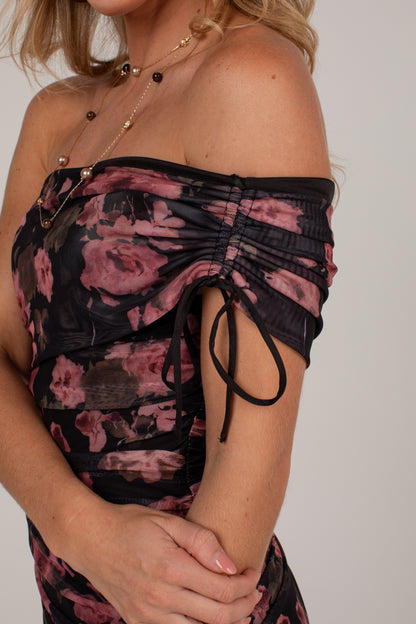 Close-up of a woman wearing an off-the-shoulder ruched top with a black and dusty pink floral print, paired with layered gold necklaces, against a plain light gray background.