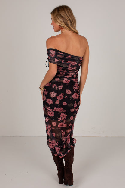 A woman shown from the back wearing a black off-the-shoulder midi dress with a pink floral print and dark brown knee-high boots, set against a plain light gray studio background.