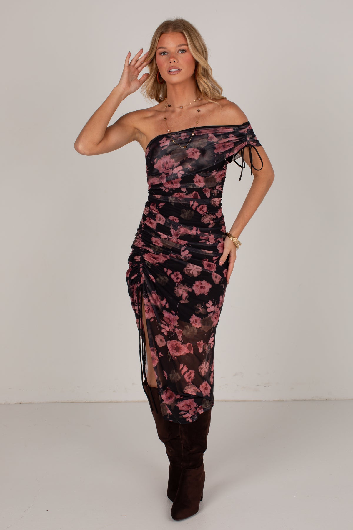 A woman models a one-shoulder ruched maxi dress with a pink and black floral pattern, styled with dark brown suede boots against a plain light gray studio background.