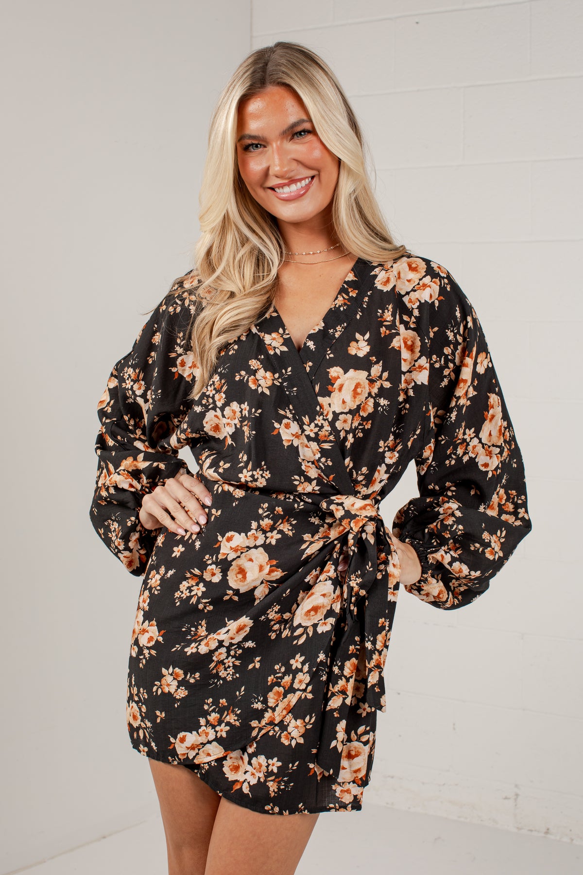A smiling blonde woman models a black wrap-style mini dress with a warm-toned floral print in orange, beige, and soft pink. The dress features long balloon sleeves and a self-tie belt at the waist, set against a clean white paneled wall background.