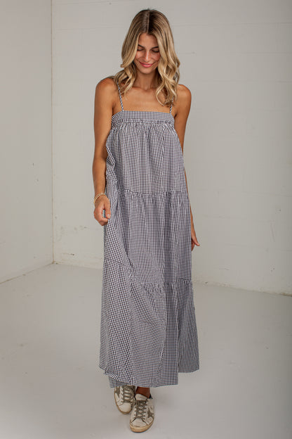 A woman modeling a sleeveless, tiered maxi dress with a small black and white gingham pattern, standing against a plain white studio wall.