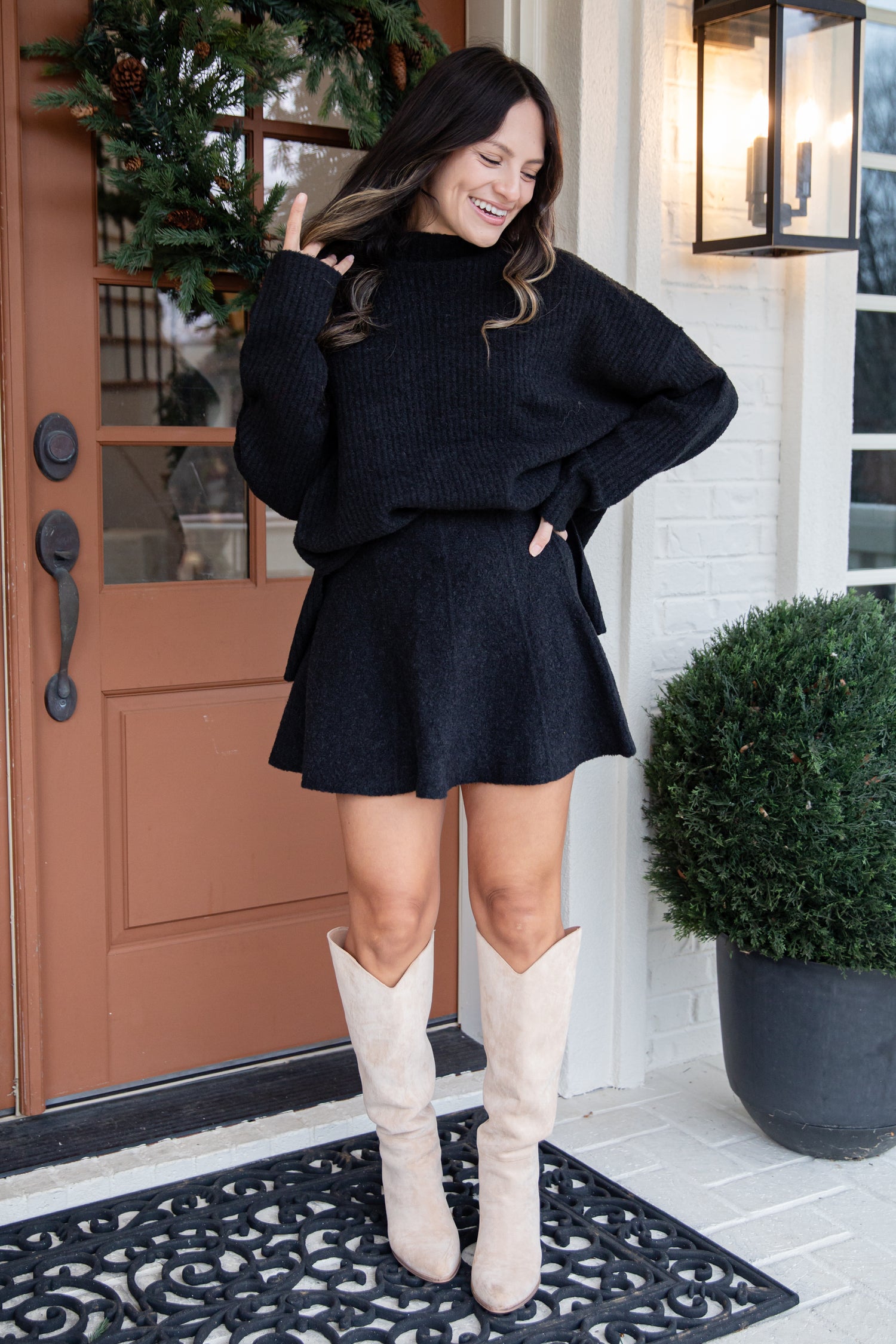 Woman in a black outfit with beige boots standing in front of a door.