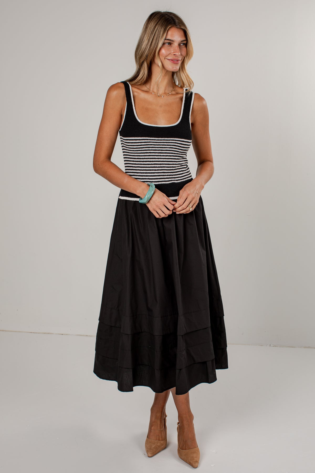 A woman modeling a sleeveless midi dress with a black and white horizontally striped bodice and a solid black tiered skirt, paired with tan pointed-toe heels against a plain light gray studio background.