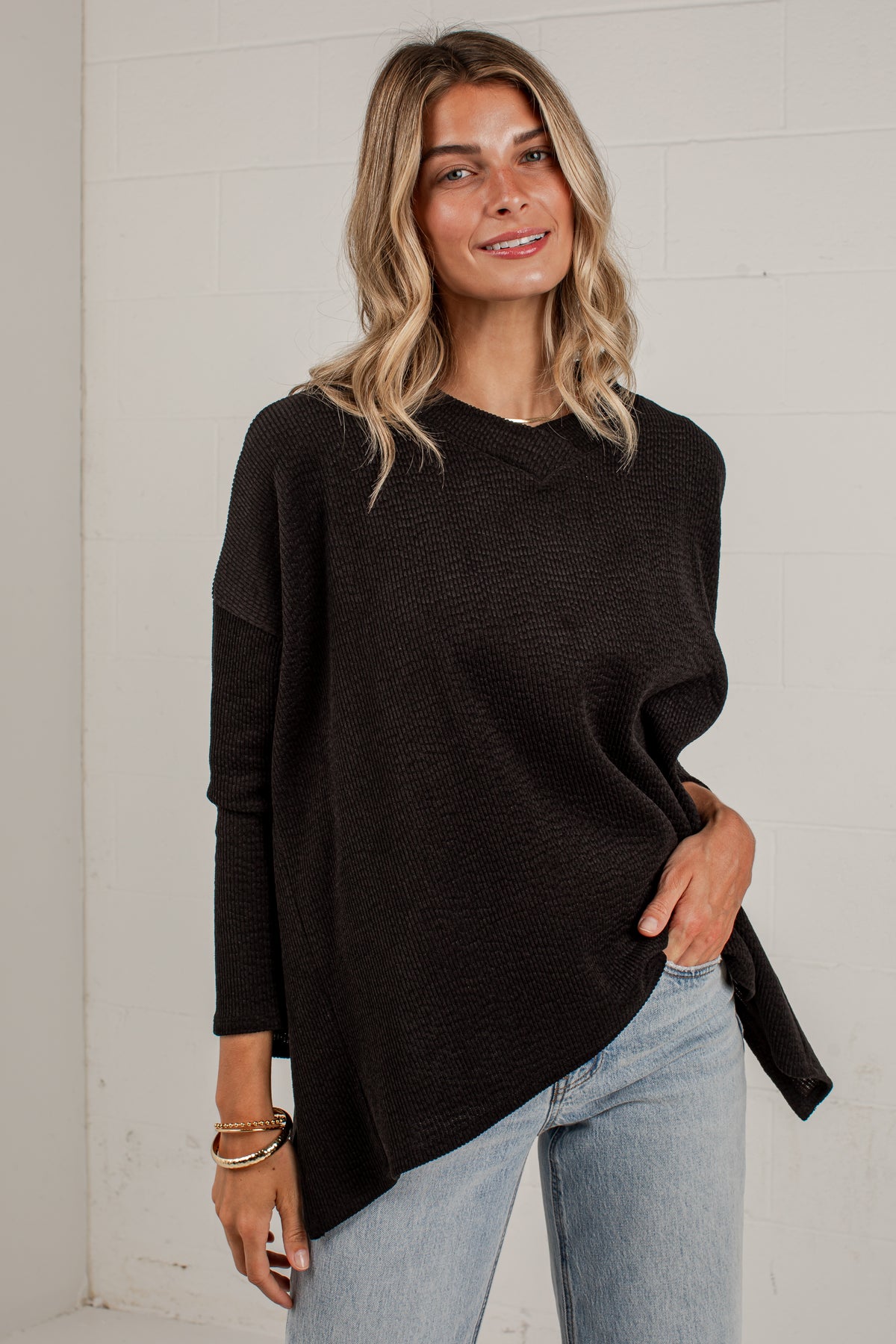 A woman wearing a black long-sleeve knit sweater with an asymmetrical hem and light-wash blue jeans, standing against a white brick wall.