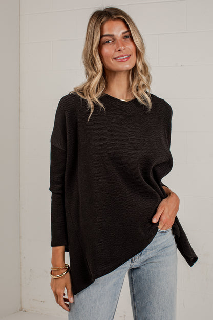 A woman wearing a black long-sleeve knit sweater with an asymmetrical hem and light-wash blue jeans, standing against a white brick wall.