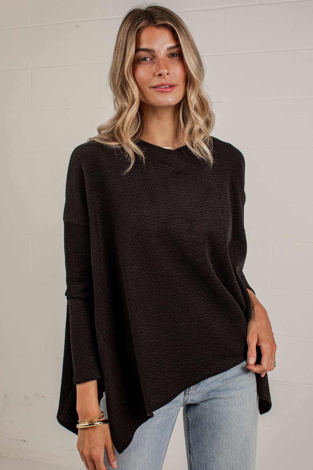 A woman wearing a dark brown long-sleeve knit top with a relaxed, slightly asymmetrical hem, paired with light blue jeans, standing against a white brick wall.
