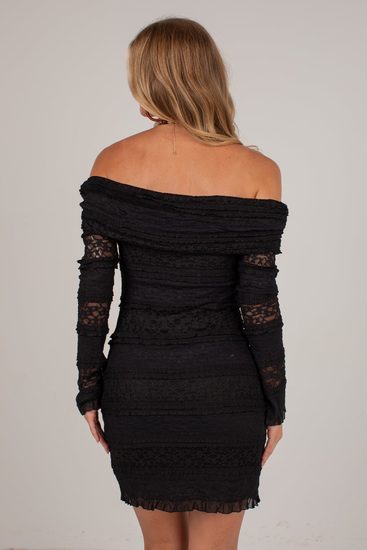 A woman shown from the back wearing a black off-the-shoulder mini dress with long sheer lace sleeves, photographed against a plain light gray studio background.