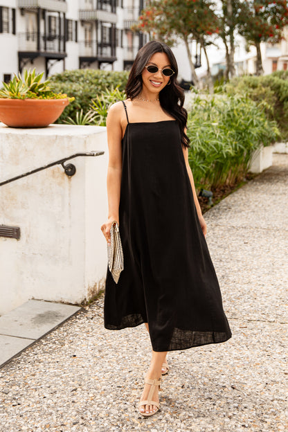 Woman in a black dress standing on a sidewalk with greenery and buildings in the background