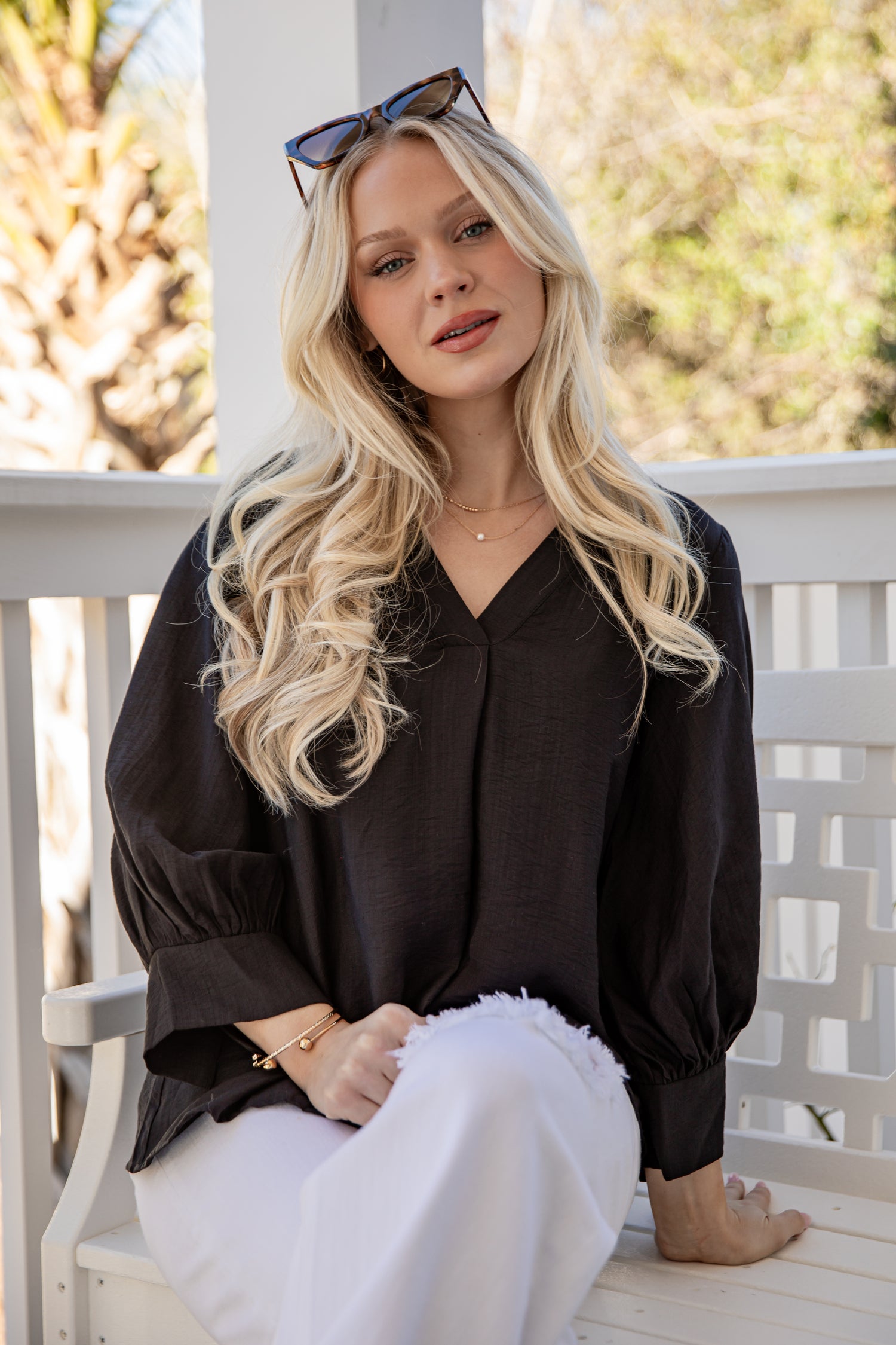 Woman with long blonde hair wearing a black top and white pants sitting on a wooden bench.
