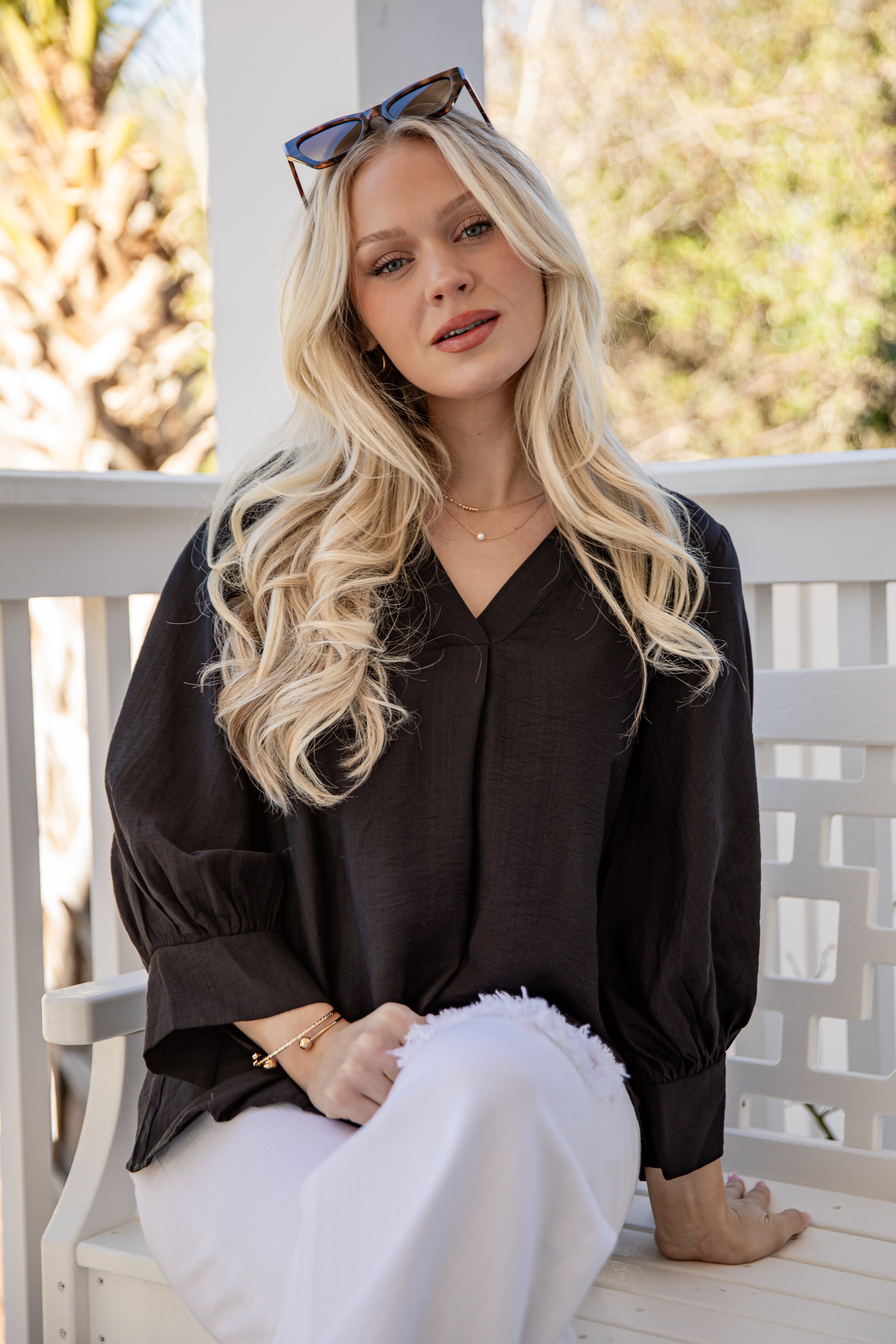 Woman with long blonde hair wearing a black top and white pants sitting on a wooden bench.