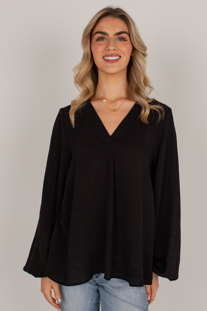 A woman with blonde wavy hair wearing a black long-sleeve V-neck top and light blue jeans, standing against a solid light gray studio background.