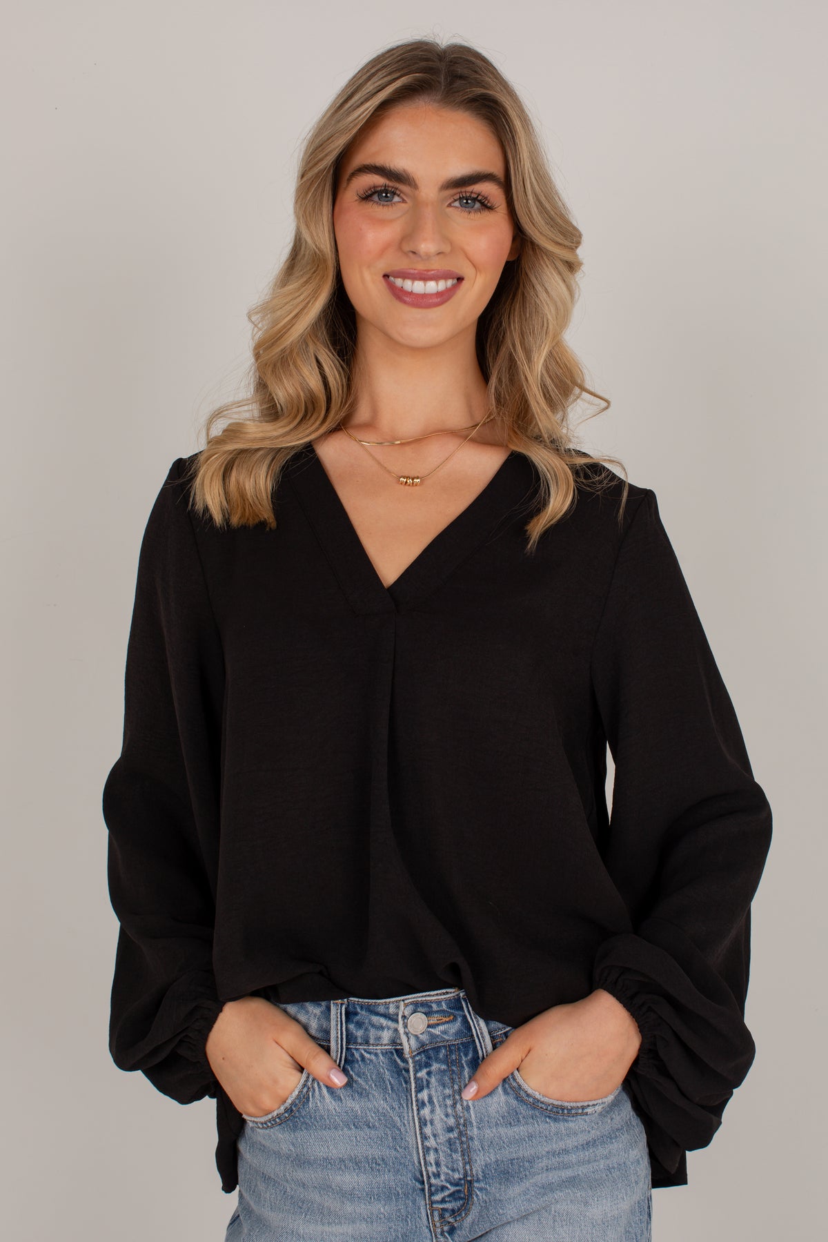 A woman with blonde hair wearing a black long-sleeve blouse with a V-neck and gathered cuffs, paired with light blue jeans, standing against a neutral gray background.
