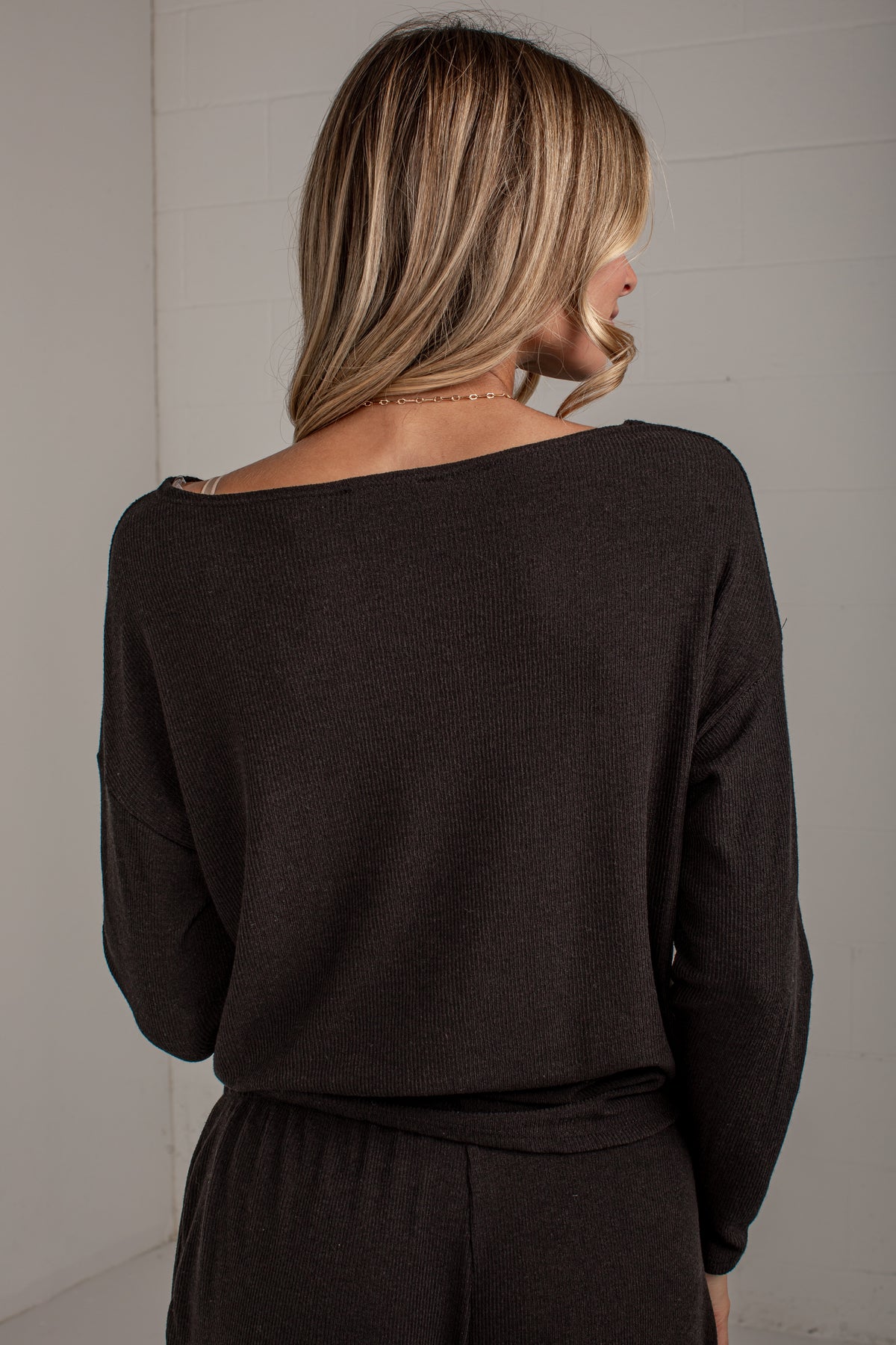 A woman viewed from behind wearing a dark brown off-the-shoulder knit sweater and matching dark bottoms, standing against a light gray paneled wall.