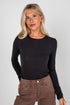 Woman wearing a black long-sleeve top and brown pants against a plain background