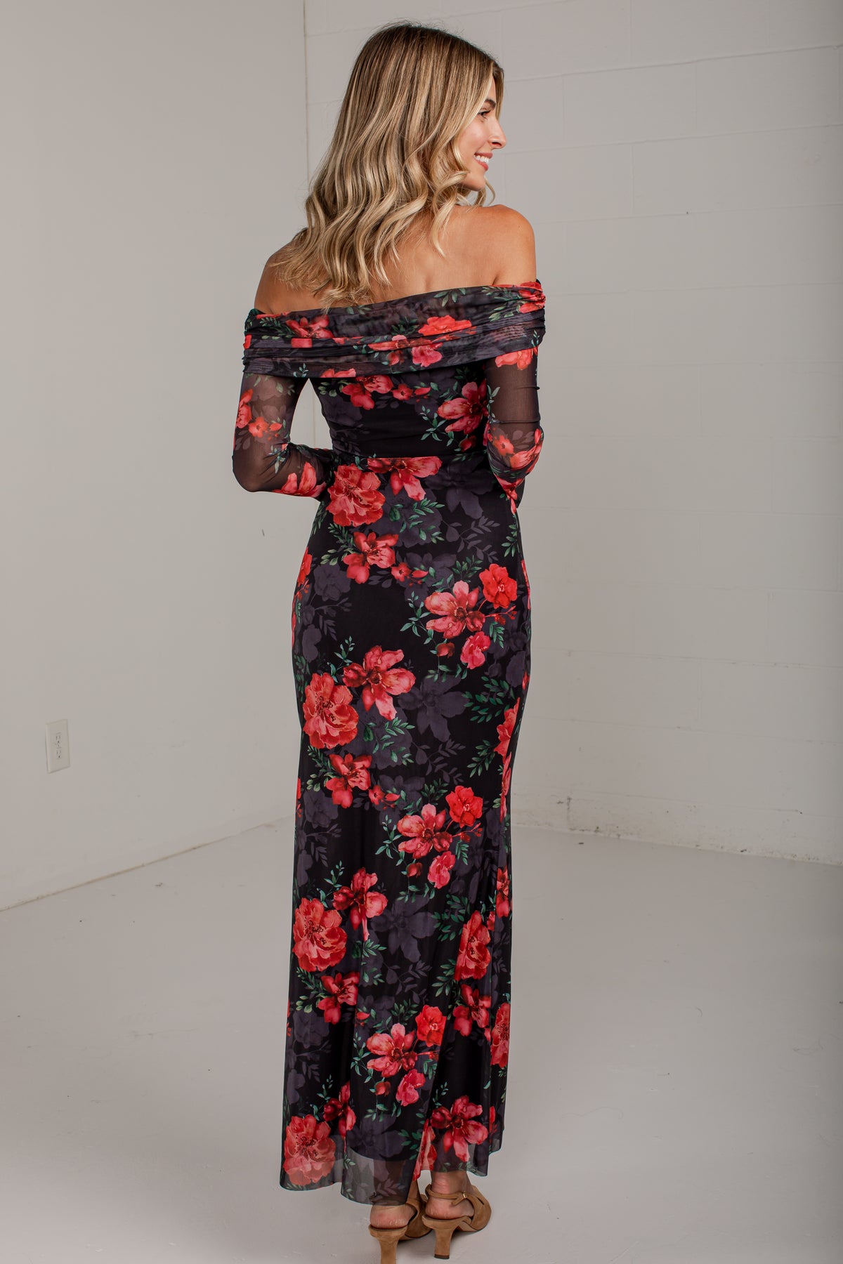 A woman wearing an off-the-shoulder black maxi dress with a vibrant red and pink floral print, photographed from the back in a minimalist white studio setting.