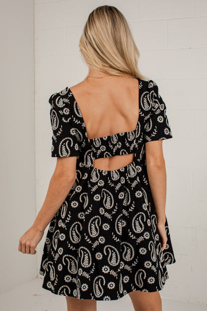 A woman viewed from the back wearing a short black dress with a white paisley and floral print, featuring puff sleeves and a cutout at the back, set against a white brick wall.