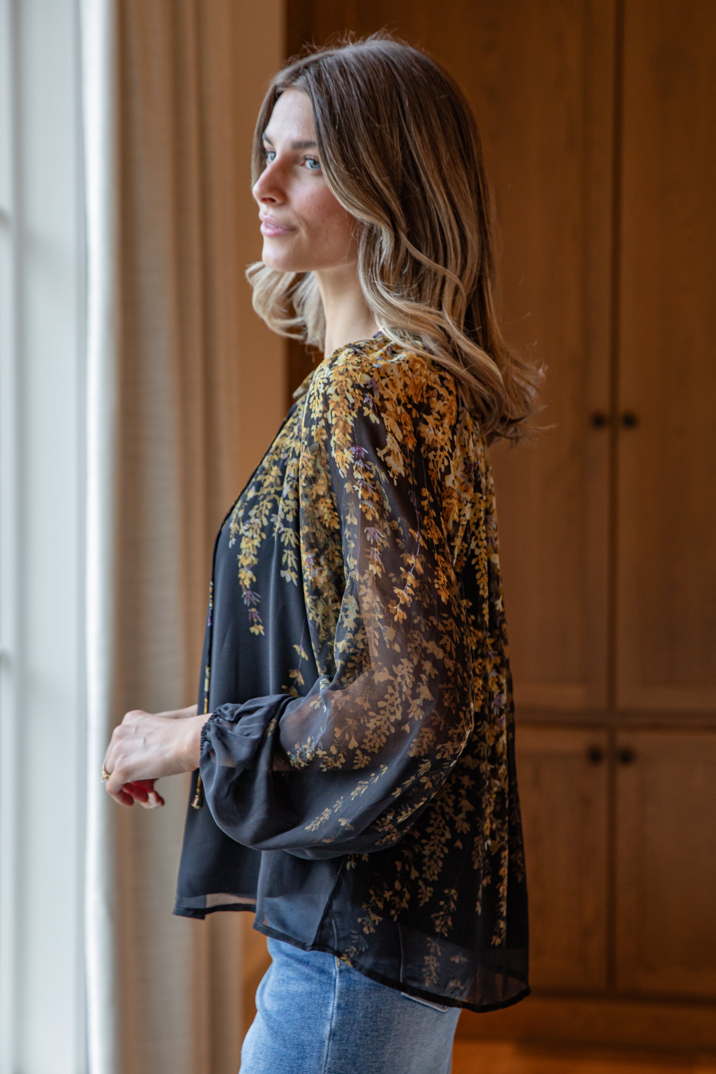 Woman wearing a floral blouse and jeans standing by a window.