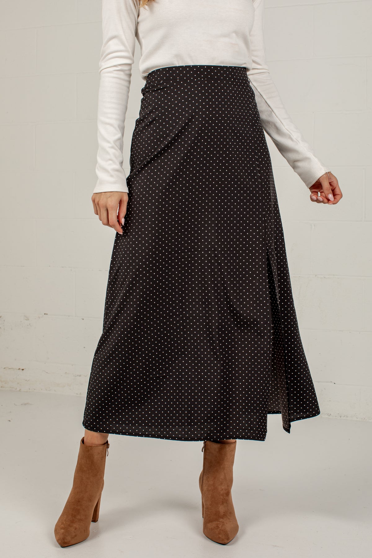A woman wearing a black midi skirt with a small white polka dot pattern, styled with a white long-sleeve top and brown suede ankle boots against a white brick wall.