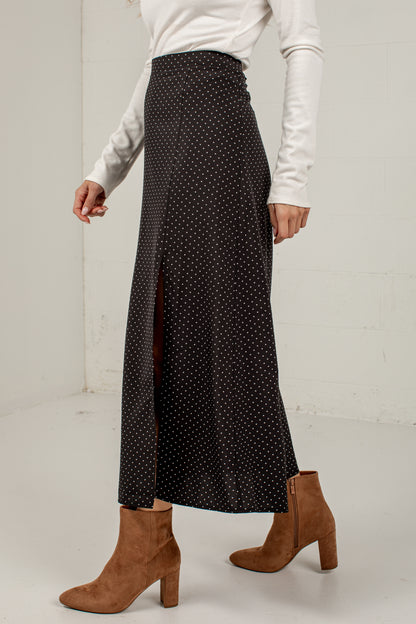 A model wearing a black midi skirt with a white polka dot pattern and a front slit, styled with a white long-sleeve top and brown suede ankle boots.