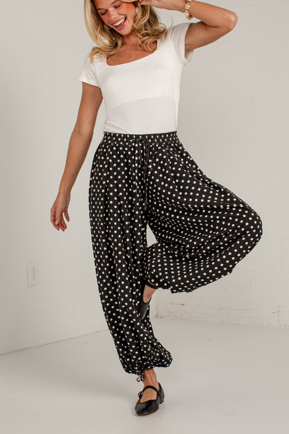 A woman modeling black wide-leg pants with white polka dots, paired with a white scoop-neck top and black flats, posing in a bright white room.