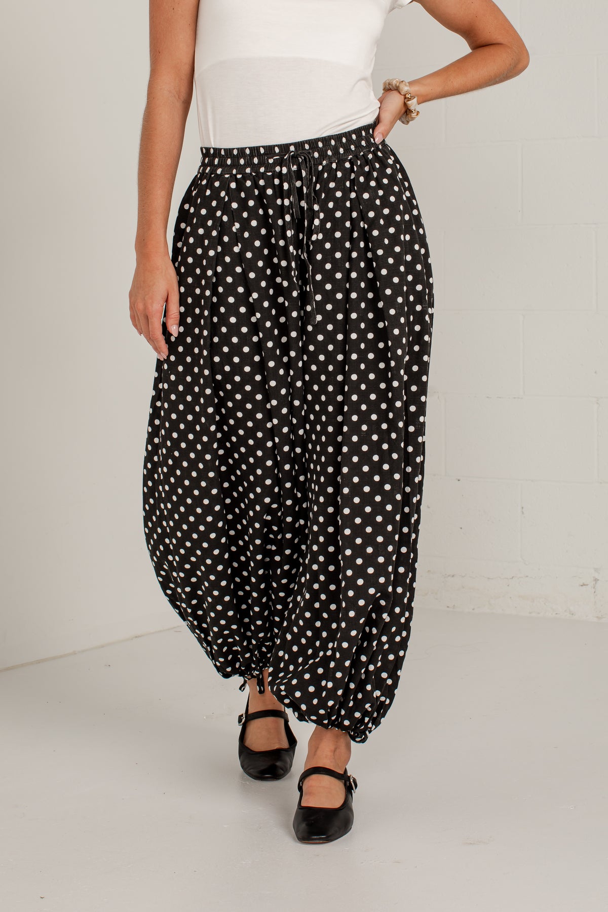 A person wearing loose black harem-style pants with a white polka dot pattern, paired with a white top and black Mary Jane shoes, standing against a white wall.