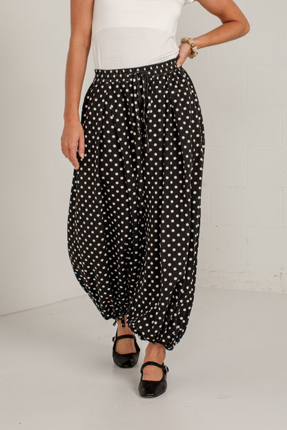 A person wearing loose black harem-style pants with a white polka dot pattern, paired with a white top and black Mary Jane shoes, standing against a white wall.