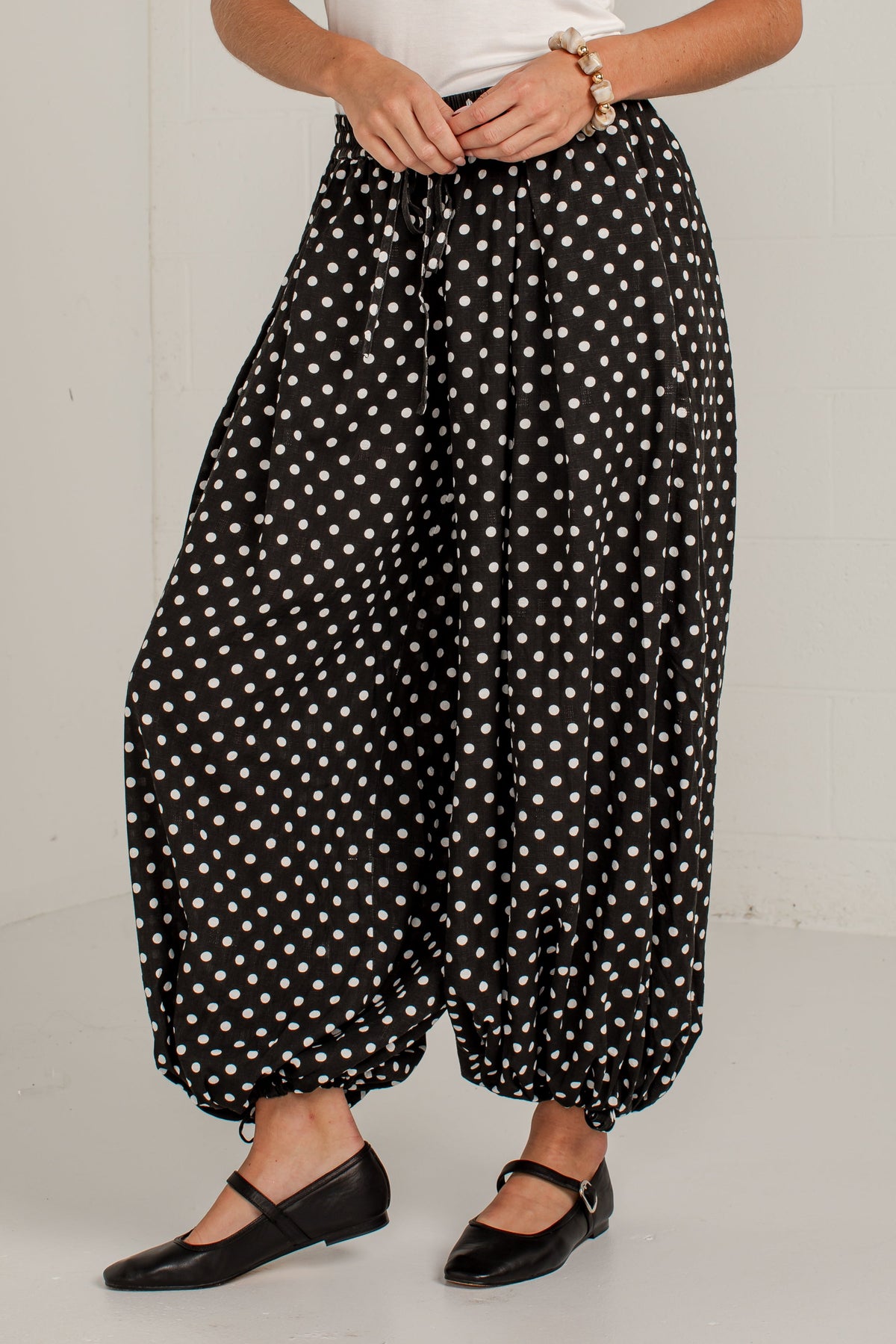 A person wearing loose black harem-style pants with white polka dots, paired with a white top and black flats, standing against a plain light gray wall.